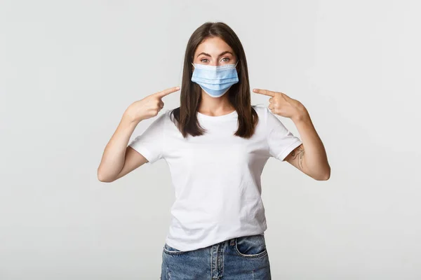 Covid-19, Gesundheits- und Sozialdistanzierungs-Konzept. Attraktives brünettes Mädchen in medizinischer Maske zeigt mit dem Finger auf Gesicht, weißer Hintergrund — Stockfoto