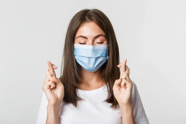 Covid-19, gezondheid en sociale afstand concept. Close-up van hoopvol mooi meisje in het gezicht masker sluit ogen en kruis vingers veel geluk, het maken van wens — Stockfoto