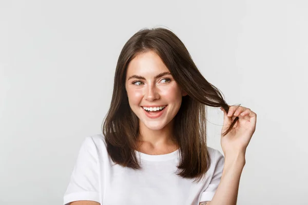 Primer plano de coqueta chica morena atractiva jugando con hebra de pelo, sonriendo y mirando intrigado a la derecha —  Fotos de Stock