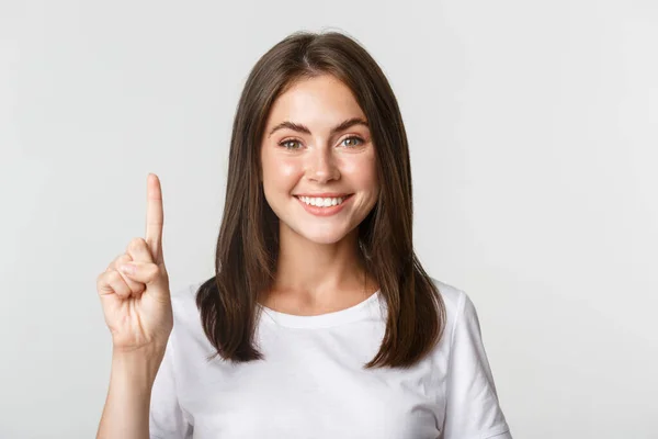 Nahaufnahme einer fröhlichen attraktiven jungen Frau, die lächelt und einen Finger zeigt, weißer Hintergrund — Stockfoto