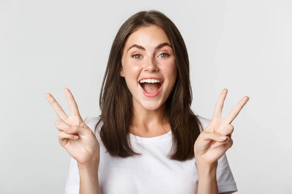 Retrato de hermosa y feliz chica morena sonriendo y mostrando gestos de paz, de pie fondo blanco emocionado — Foto de Stock