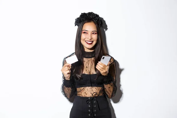 ゴシックレースのドレスと黒の花輪で美しいアジアの女性のイメージ,脇に見て満足し、笑顔,クレジットカードで携帯電話を保持 — ストック写真