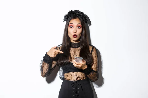 Retrato de chica asiática asombrada apuntando con el dedo al teléfono inteligente, mostrando algo sobre halloween en internet, de pie sobre fondo blanco en vestido de encaje gótico — Foto de Stock