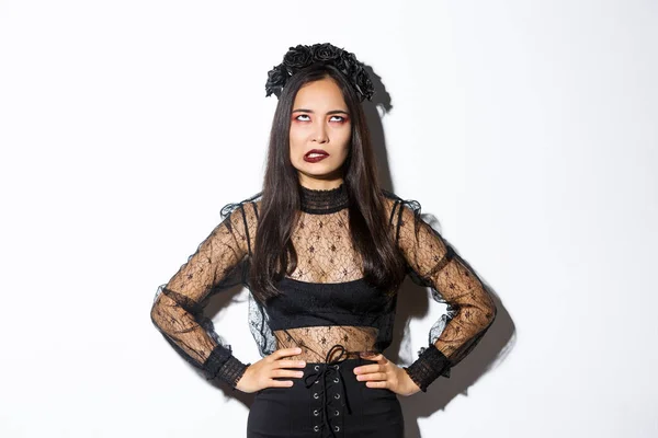 Annoyed asian woman in witch costume rolling eyes and looking pressured. Irritated girl hate celebrating halloween, looking pissed-off, wearing gothic black lace dress and wreath — стоковое фото