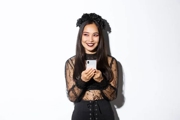 Retrato de mujer asiática hermosa astuta, bruja en vestido de encaje gótico usando el teléfono móvil, sonriendo y mirando a la esquina superior izquierda, de pie sobre fondo blanco — Foto de Stock