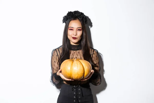Afbeelding van sluwe aziatische vrouw in zwarte jurk, imiteren kwade heks op Halloween, met grote pompoen, staande over witte achtergrond — Stockfoto
