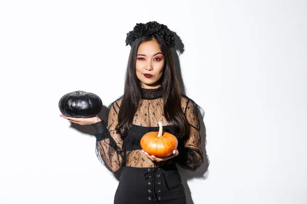 Descarada atractiva chica asiática en traje de bruja, la celebración de calabazas y mirando a la cámara, de pie sobre fondo blanco —  Fotos de Stock