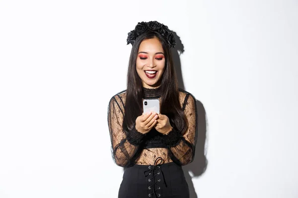 Eccitato donna sorridente guardando il telefono cellulare soddisfatto, indossando abito in pizzo gotico per la festa di Halloween, in piedi su sfondo bianco — Foto Stock
