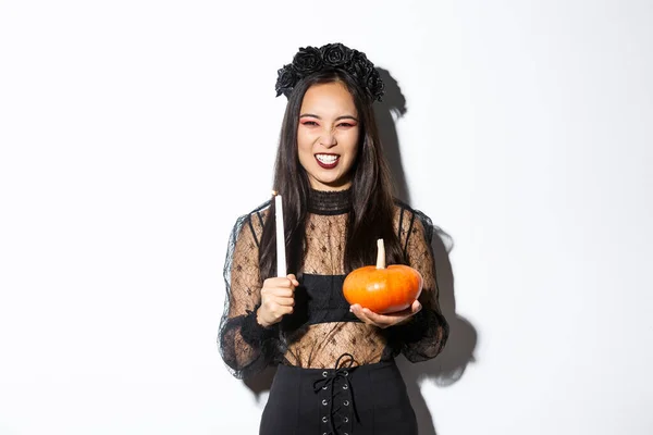 Image of asian evil witch in gothic lace dress and black wreath, laughing and grimacing, holding candle with pumpkin, celebrating halloween — Stock Photo, Image