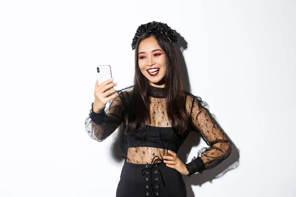 Ritratto di felice bella donna asiatica in costume di Halloween sorridente e guardando lo schermo del telefono cellulare, avendo videochiamata, in piedi su sfondo bianco — Foto Stock