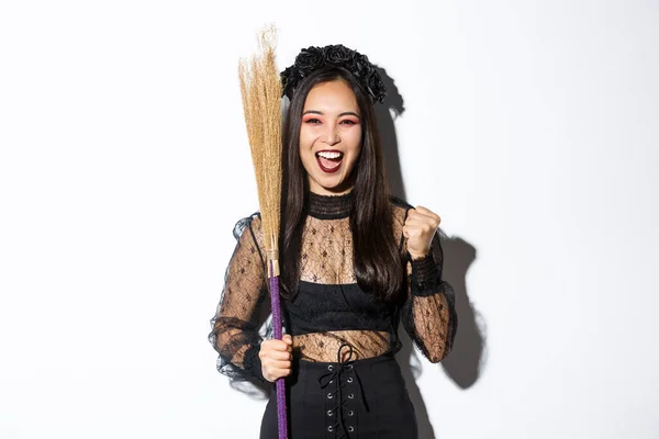 Imagen de chica asiática alegre en traje de bruja celebrando la victoria, sosteniendo la escoba, diciendo sí y levantando el puño en triunfo, fondo blanco —  Fotos de Stock