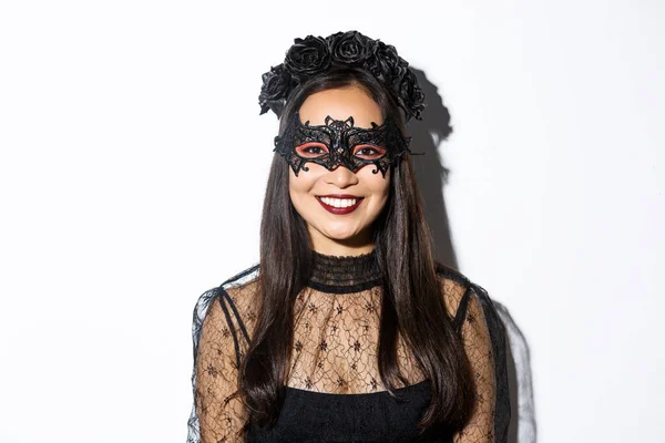 Крупный план таинственной женщины в готическом венке и черной маске, улыбающейся в камеру, празднующей Хэллоуин, стоящей на белом фоне — стоковое фото