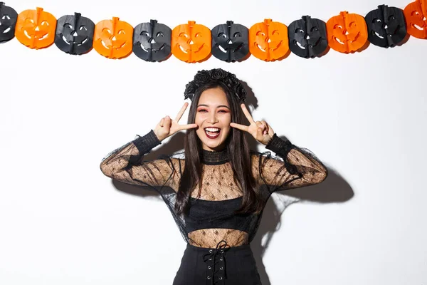 Carino felice donna asiatica godendo festa di Halloween, mostrando gesto di pace e sorridente, indossando costume da strega, in piedi contro la decorazione banner zucca — Foto Stock