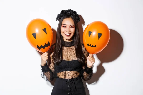 Imagem de mulher asiática alegre em traje de bruxa celebrando halloween, segurando balões com rostos assustadores, de pé sobre fundo branco — Fotografia de Stock