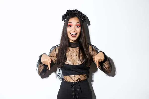 Glücklich schöne asiatische Frau in Halloween-Gothic-Kleid, zeigt mit dem Finger nach unten als Anzeige, zeigt Ihre Promo, steht über weißem Hintergrund — Stockfoto