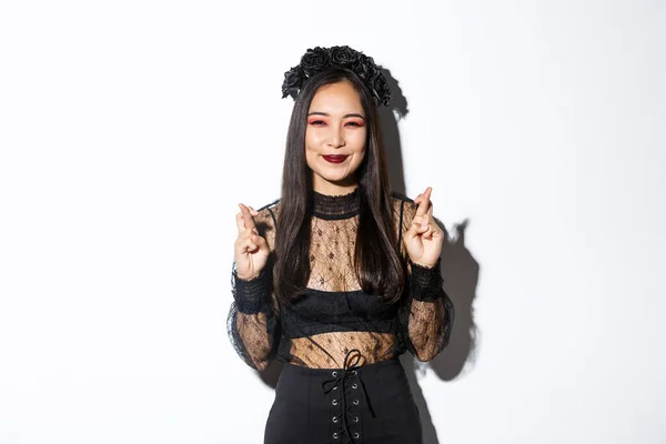 Imagen de la feliz chica asiática que le encanta celebrar Halloween disfrazado de bruja, sonriendo esperanzado y cruzar los dedos para la buena suerte, pidiendo deseo, de pie sobre fondo blanco —  Fotos de Stock