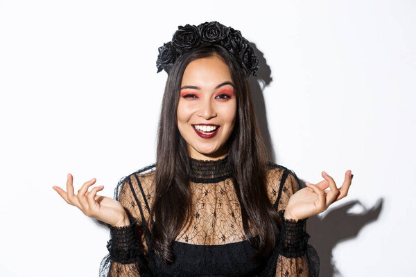 Close-up of sassy pretty asian woman in wicked witch costume making evil laugh and spread hands sideways, standing over white background