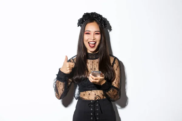 Retrato de mulher asiática satisfeita em vestido gótico elegante e grinalda preta mostrando polegares para cima ao usar o telefone móvel, de pé sobre fundo branco — Fotografia de Stock