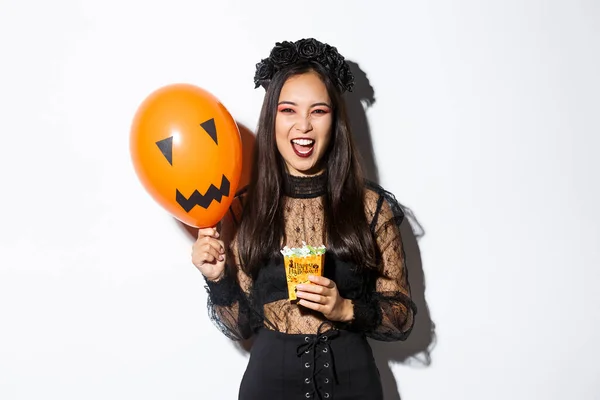 Enge heks truc of traktatie op Halloween, met snoep en oranje ballon, staande in gotische kant jurk met zwarte krans — Stockfoto