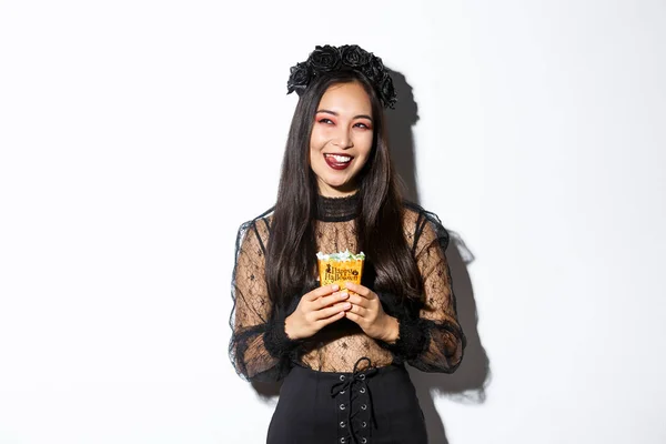 Vrolijke mooie aziatische vrouw vieren Halloween in heks kostuum, met snoep en glimlachen, kijken naar de linker bovenhoek sluwe — Stockfoto