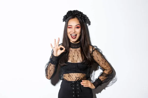 Aantrekkelijke jonge aziatische vrouw in halloween gotische jurk, tonen oke gebaar, toestemming geven, goedkeuren of graag uw feest kostuum, staande blij over witte achtergrond — Stockfoto