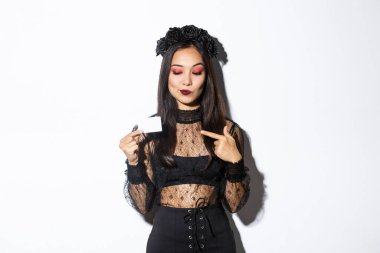 Cadı kostümlü güzel Asyalı kızın resmi kredi kartını işaret ediyor, beyaz arka planda duruyor.