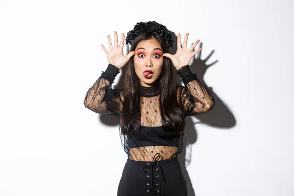 Retrato de mujer asiática divertida y tonta en traje de bruja celebrando halloween, burlándose de alguien, mostrando caras y lengua pegajosa, de pie sobre fondo blanco —  Fotos de Stock