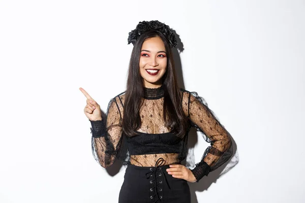 Tevreden lachende aziatische vrouw in kwade heks of banshee kostuum vieren Halloween, op zoek blij en wijzende vinger linker bovenhoek, het tonen van uw promo banner, witte achtergrond — Stockfoto