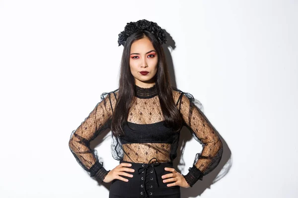 Attraente donna asiatica in costume di Halloween cercando deluso e scettico. Donna in pizzo nero vestito e ghirlanda cercando arrogante, dolcetto o scherzetto in abito da strega, in piedi sfondo bianco — Foto Stock