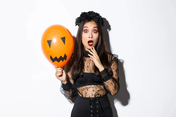 Retrato de mujer asiática sorprendida en traje de halloween, disfrazada de bruja, sosteniendo globo naranja con cara aterradora — Foto de Stock