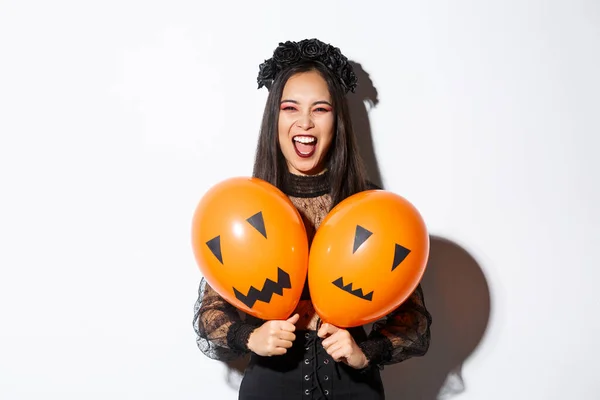 Afbeelding van aziatische meisje in kwade heks kostuum met twee oranje ballonnen met enge gezichten, vieren Halloween, staande over witte achtergrond — Stockfoto