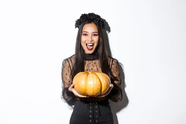Imagem de mulher asiática alegre em traje de bruxa, celebrando halloween, dando-lhe abóbora e olhando assustador, de pé sobre fundo branco — Fotografia de Stock