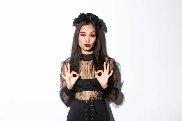 Image of stylish asian woman in gothic dress, looking satisfied and showing okay gestures in approval, praise something good, standing impressed over white background — Stock Photo, Image