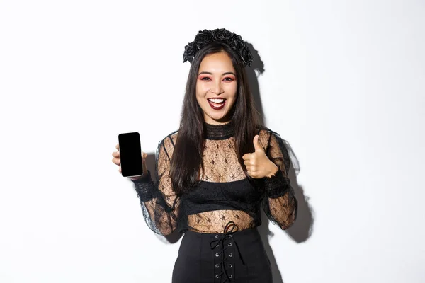 Eccitato e soddisfatto donna asiatica in costume di Halloween mostrando pollici-up in approvazione e dimostrare schermo del telefono cellulare, in piedi su sfondo bianco — Foto Stock