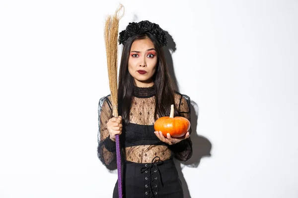 Endişeli ve kafası karışmış cadı kostümlü Asyalı kadın gergin görünüyor, elinde süpürge ve balkabağı, cadılar bayramında şeker mi şaka mı, beyaz arka planda duruyor. — Stok fotoğraf