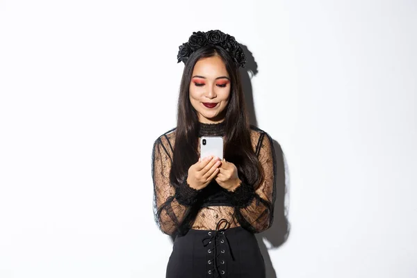 Immagine di elegante donna asiatica in costume di Halloween controllare i messaggi sul telefono cellulare. Ragazza in abito di pizzo gotico guardando smartphone, in piedi su sfondo bianco — Foto Stock