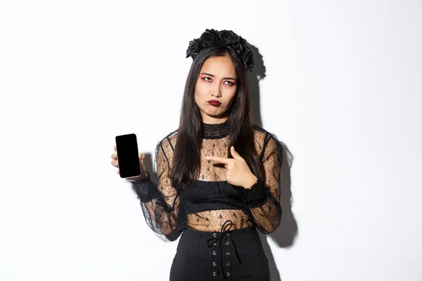 Imagem de menina asiática irritada e relutante em traje de bruxa, rolar os olhos e amuar ao apontar o dedo para a tela do telefone móvel, de pé sobre fundo branco — Fotografia de Stock