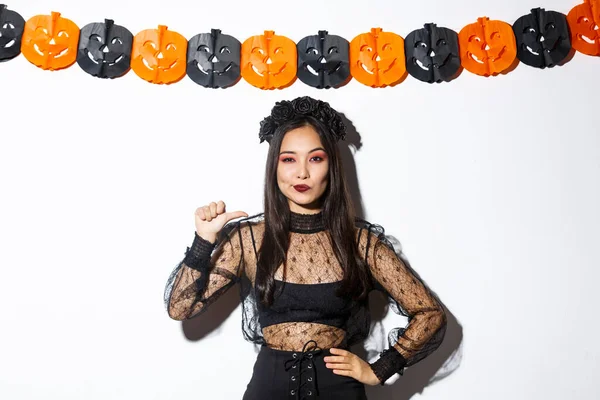 Fiduciosa donna asiatica in elegante abito gotico e ghirlanda nera che punta a se stessa come professionista, in piedi contro le decorazioni di Halloween — Foto Stock