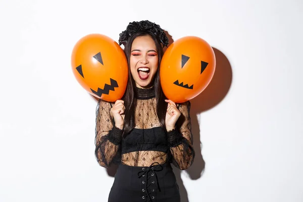 Imagine a unei fete asiatice în costum de vrăjitoare malefică care deține două baloane portocalii cu fețe înfricoșătoare, sărbătorind Halloween-ul, stând pe fundal alb — Fotografie, imagine de stoc