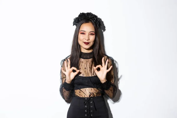 Изображение улыбающейся азиатской милой ведьмы, одетой в черно-готическое платье и венок, показывающие нормальные жесты одобрения, хвалить что-то, стоя в костюме Хэллоуина на белом фоне — стоковое фото