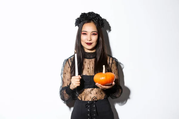 Image of devious smiling asian witch in gothic dress, holding candle with pumpkin and looking at camera cunning, standing over white background — Stock Photo, Image