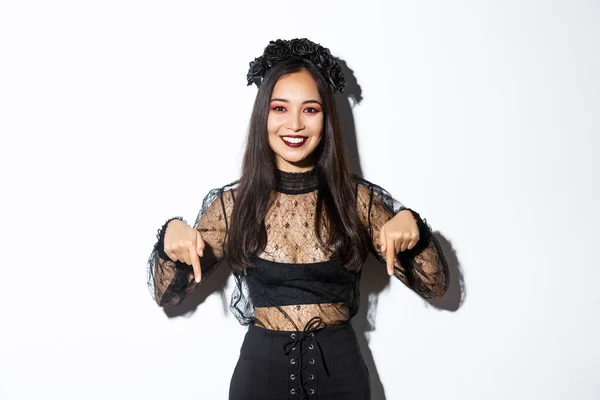 Mulher asiática bonita em traje de bruxa e maquiagem estilo gótico, apontando os dedos para baixo, mostrando banner promo sobre halloween ou dia da morte, de pé sobre fundo branco — Fotografia de Stock