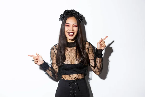 Beautiful happy young woman celebrating halloween in black lace dress, gothic style makeup, pointing fingers sideways, showing two banners or products, standing over white background — Stock Photo, Image