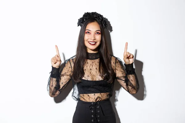 Hermosa mujer asiática en traje de viuda no muerta, vestido de encaje negro para Halloween, señalando con los dedos hacia arriba y sonriendo complacido, mirando su logotipo, de pie sobre fondo blanco —  Fotos de Stock