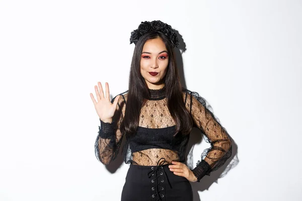 Изображение уверенной красивой азиатки в костюме Хэллоуина, показывающей пять пальцев, поднимающей руку, чтобы поздороваться, стоя на белом фоне — стоковое фото