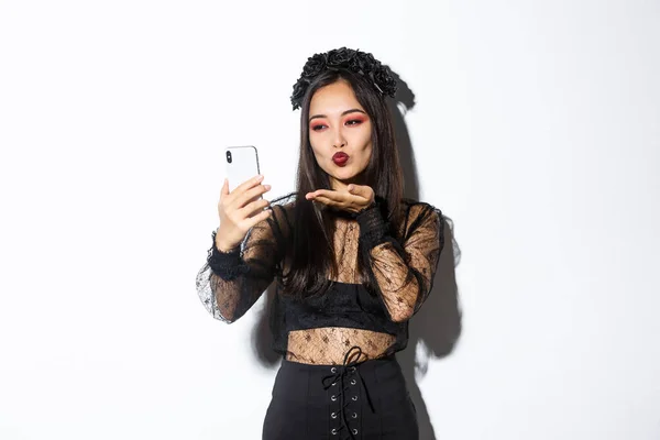 Retrato de mujer blogger asiática con estilo con maquillaje gótico y disfraz de halloween enviando un beso de aire a la cámara del teléfono móvil, grabando video o teniendo videocall, de pie sobre fondo blanco — Foto de Stock