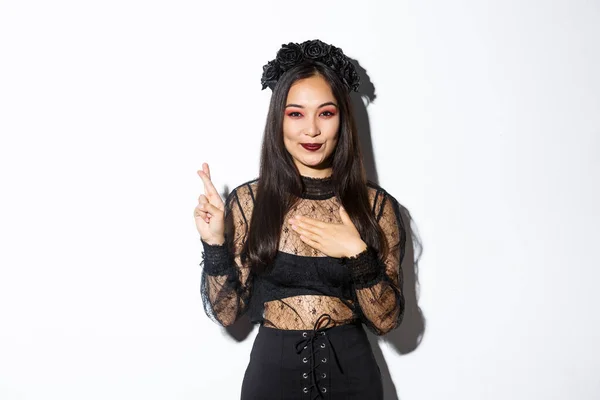 Изображение привлекательной азиатской женщины в Хэллоуин партии платье загадывая желание, держа руку на сердце и скрестив пальцы на удачу, стоя на белом фоне — стоковое фото