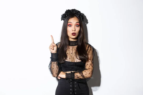 Siyah dantelli ve gotik makyajlı güzel bir Asyalı kadın düşünmüştür. Cadı kostümü giymiş kız parmağını kaldırıyor ve cadılar bayramı fikrini öneriyor. — Stok fotoğraf