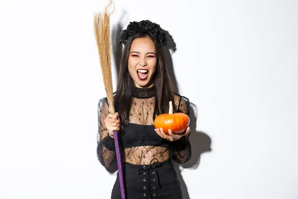 Afbeelding van mooie aziatische vrouw verkleed als een heks voor Halloween feest, met bezem en pompoen, staande over witte achtergrond — Stockfoto