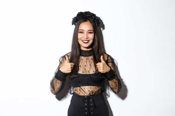 Bild av lycklig asiatisk kvinna i halloween party klänning visar tummen upp i godkännande. Flicka klädd som häxa eller änka ser nöjd, gillar eller håller med om något, vit bakgrund — Stockfoto